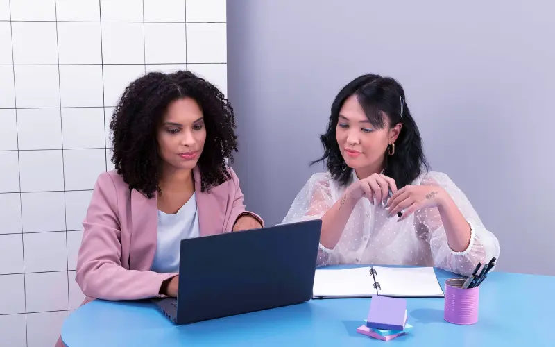 Imagem de duas mulheres em uma mesa de reunião olhando para um notebook.