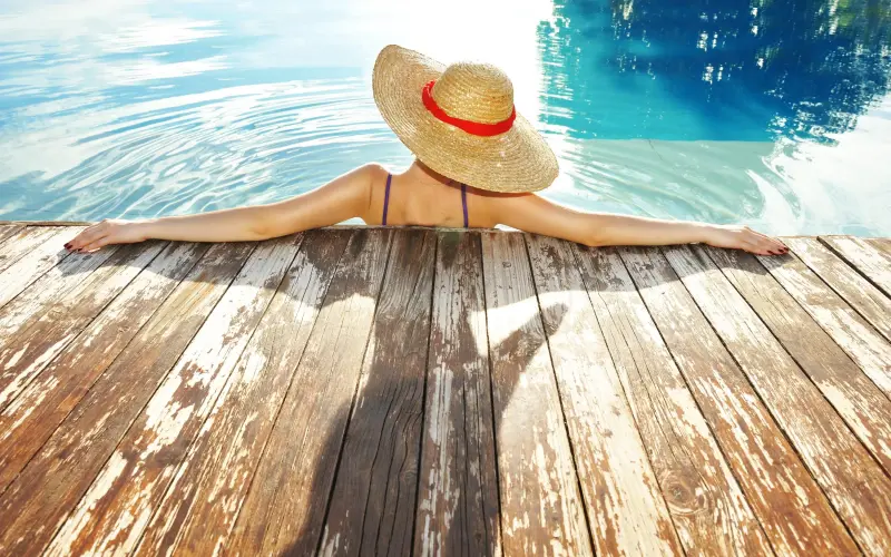 Mulher com chapéu apoiada no deck da piscina.
