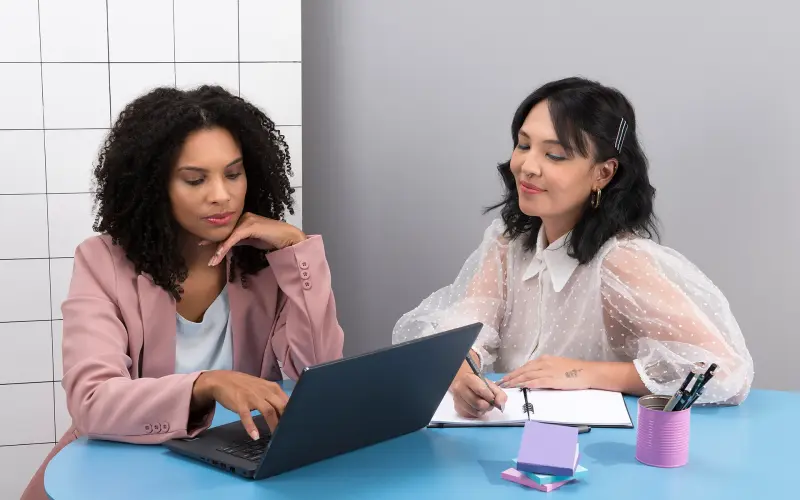 Imagem de duas mulheres sentada em frente a um notebook.