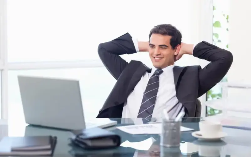 Imagem de um homem de terno sentado em frente ao seu computador, com os  braços cruzados atrás da cabeça