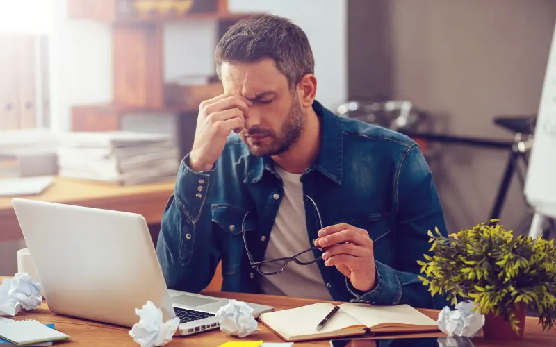 Imagem de um homem em frente ao seu computador com expressão frustrada, com diversas folhas de papel amassadas na mesa ao seu redor