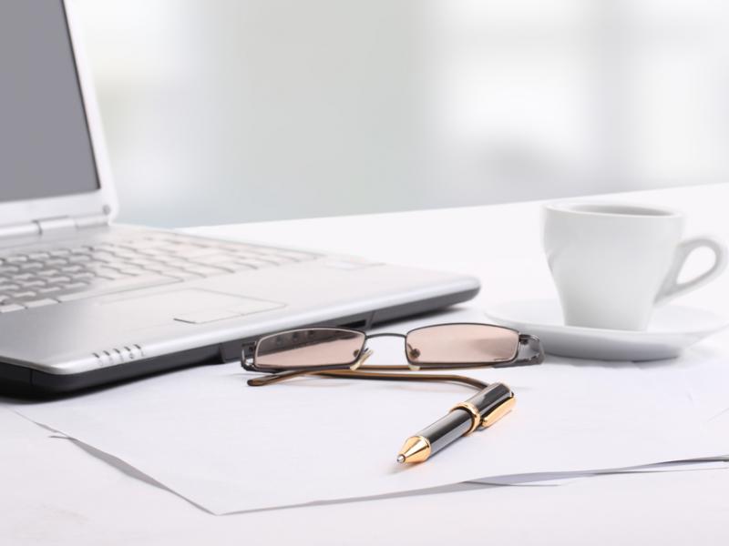 Imagem de um notebook, papéis, canetas, e uma xícara de café em cima de uma mesa branca