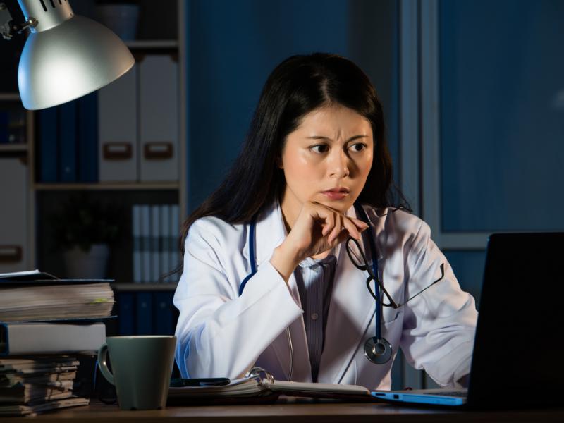 Imagem de uma mulher com jaleco de médico senta na frente de um computador em uma sala escura